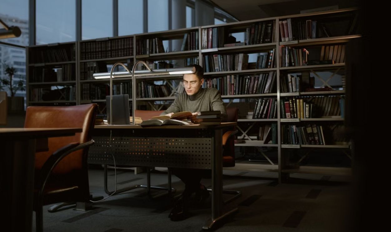 A man intensely studies in a quiet library, surrounded by books and soft lighting