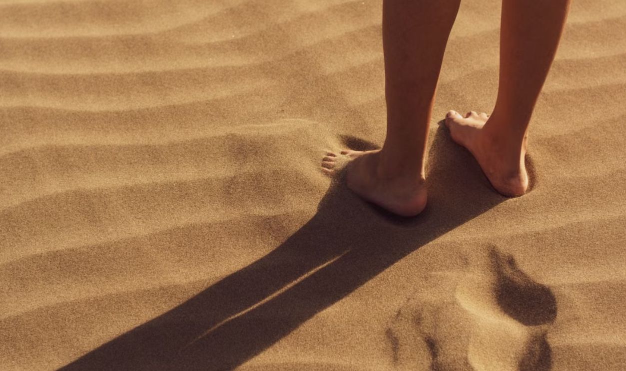 Close-up of a person standing barefoot on sunlit sand with visible footprints