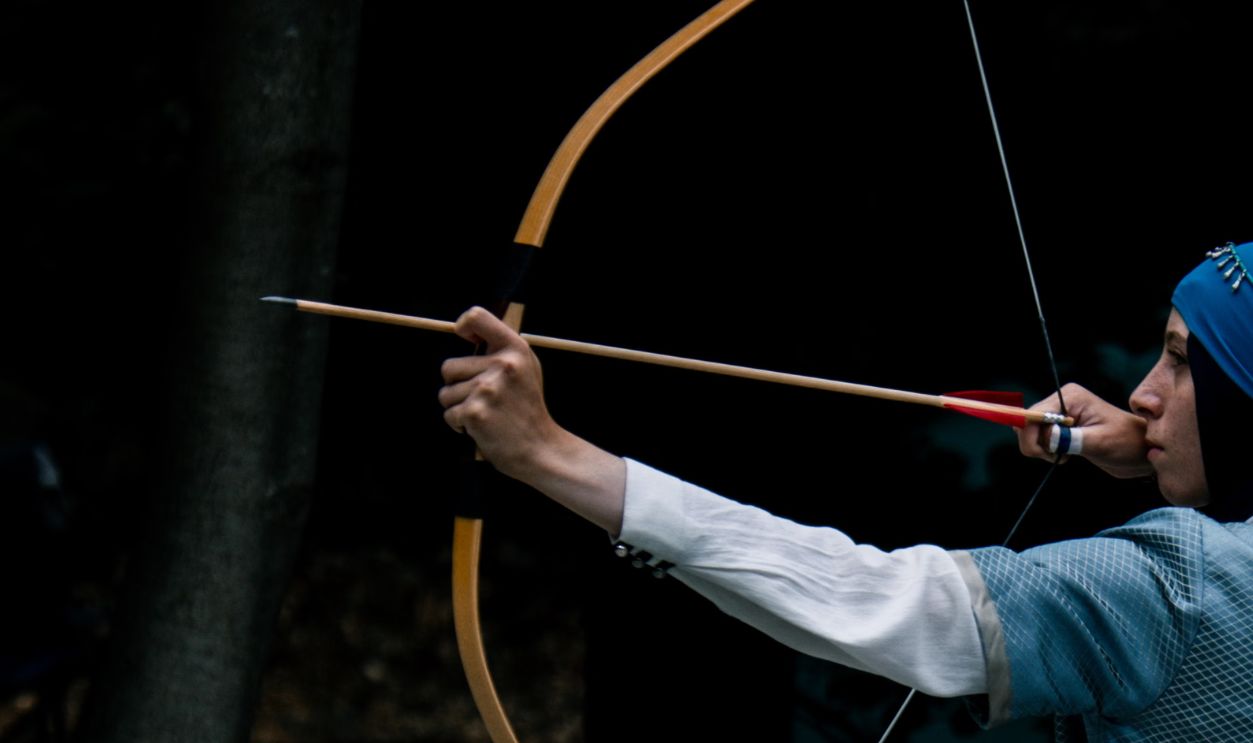 A woman in medieval costume practicing archery outdoors with a traditional bow and arrow