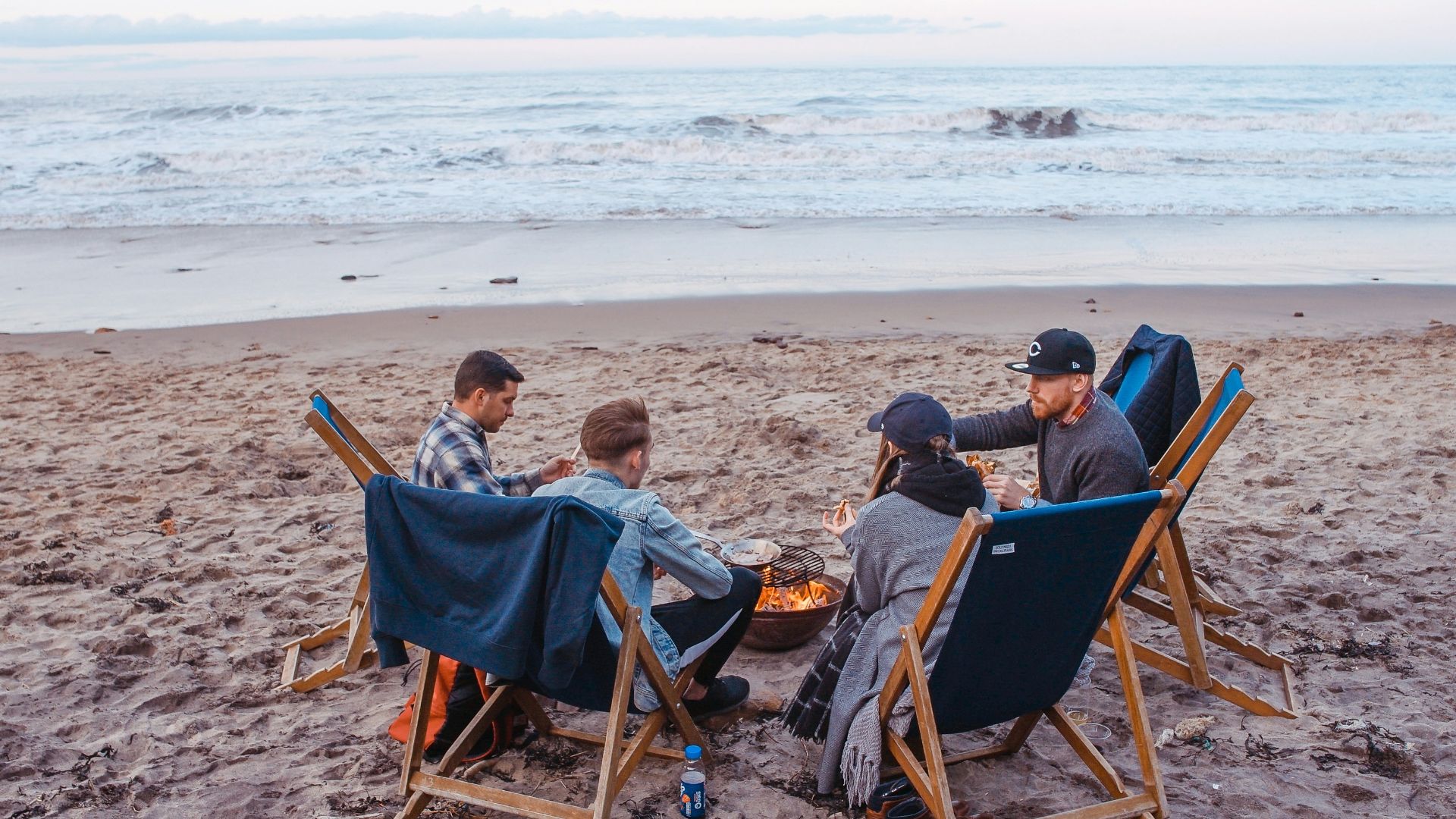 three men and one woman sitting on beach lounge in front fire pit near seashore