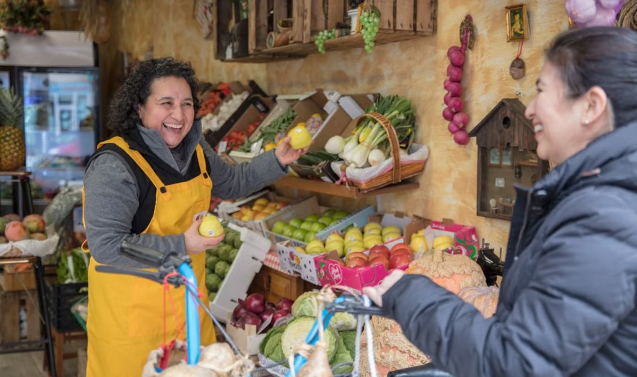 Two women smiling and exchanging fresh produce at a vibrant greengrocer's market