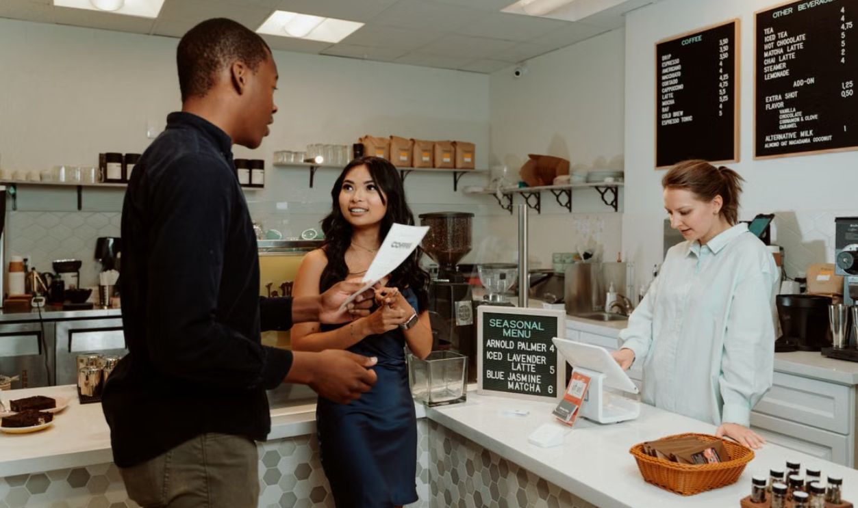 Two customers engage with a barista while ordering at a modern café counter