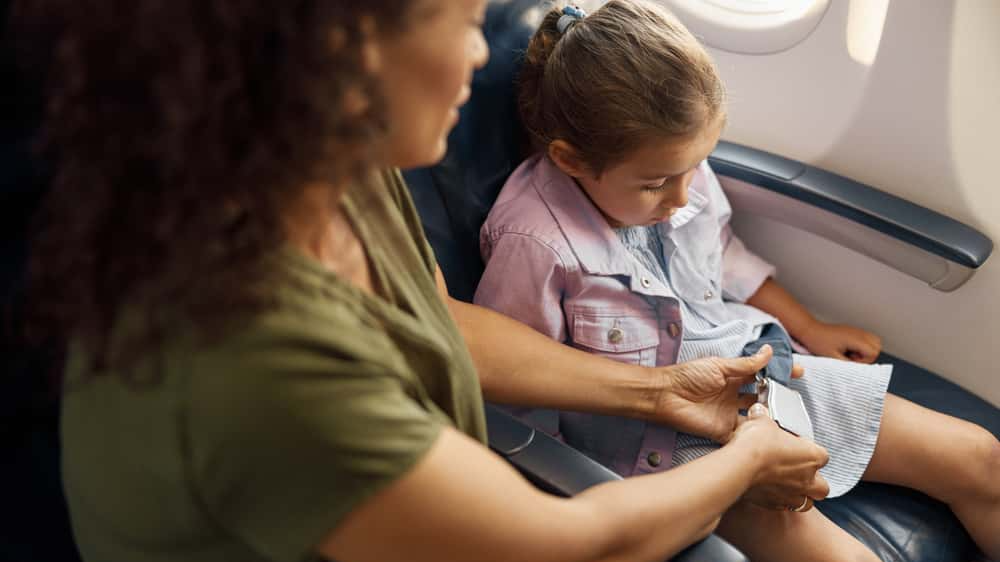 Mother helping her little daughter to adjust and tight seatbelt on an airplane for safe flight. Traveling by airplane during Covid19, family concept