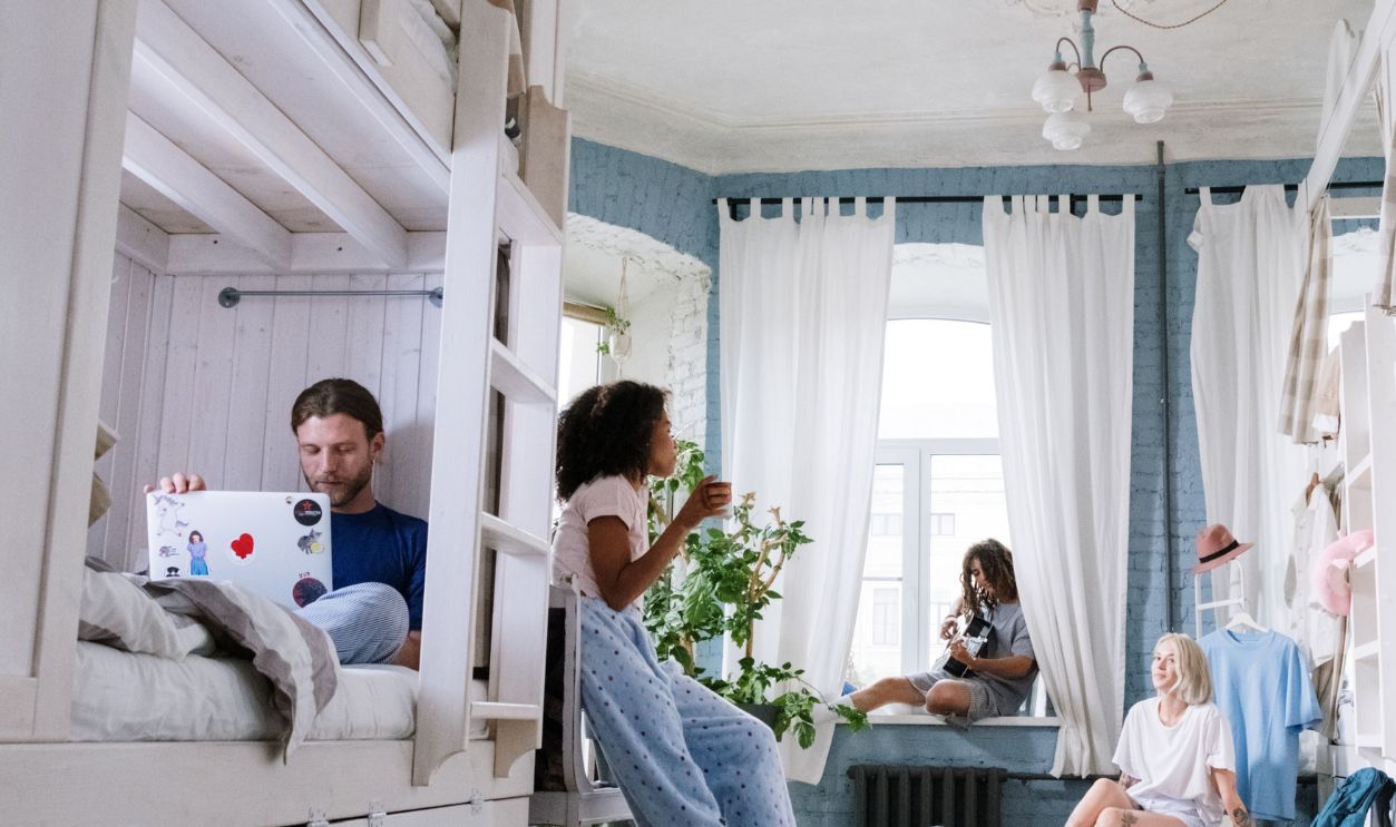 A cozy dorm room where friends relax, chat, play guitar, and use laptops together