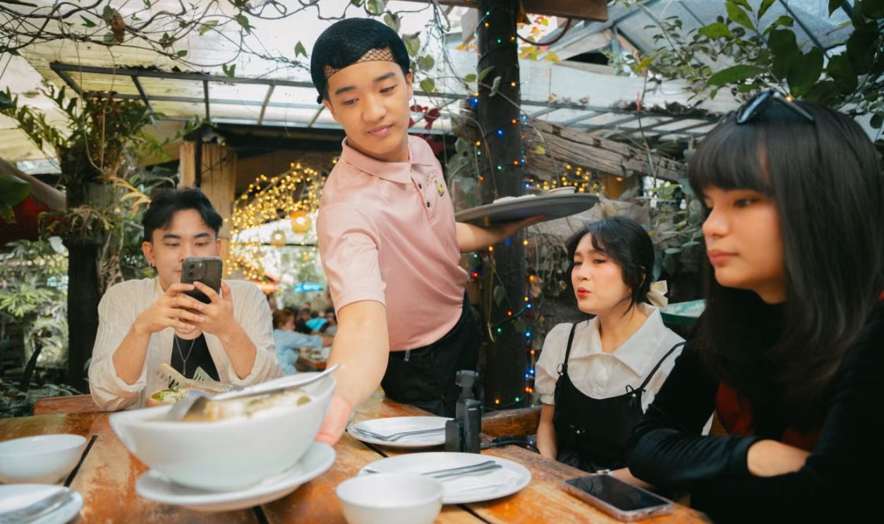 Group of friends being served by a waiter in an outdoor restaurant setting with festive lights.