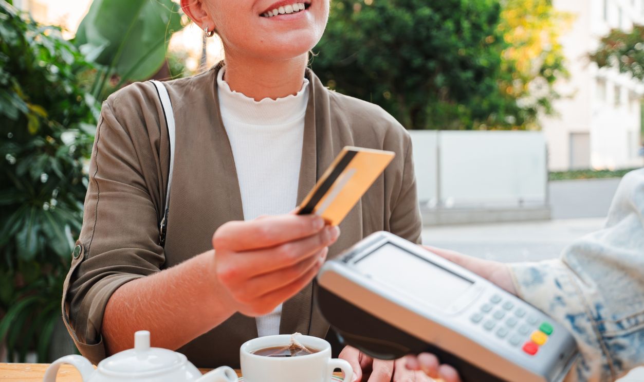 Vertical smiling young woman sitting at a bar terrace, paying for her tea using a credit card and contactless payment terminal, highlighting convenience and modern technology in outdoor dining