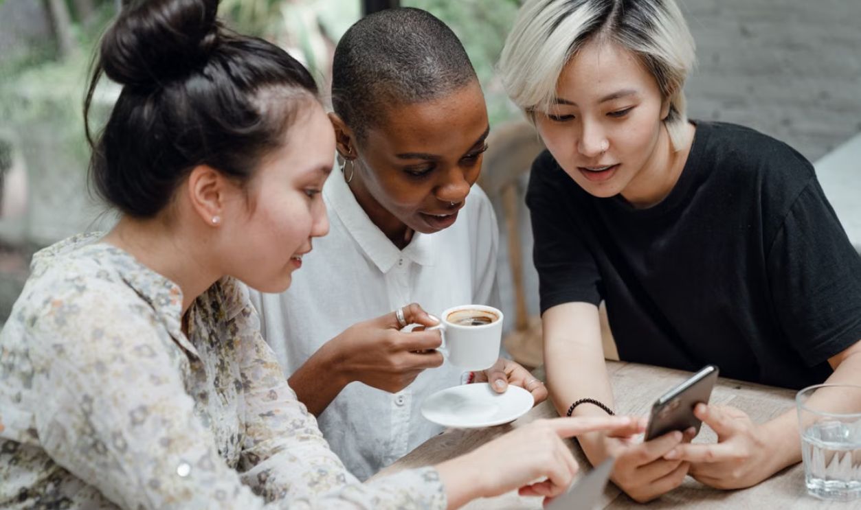 Three young women enjoying coffee and browsing social media together indoors.