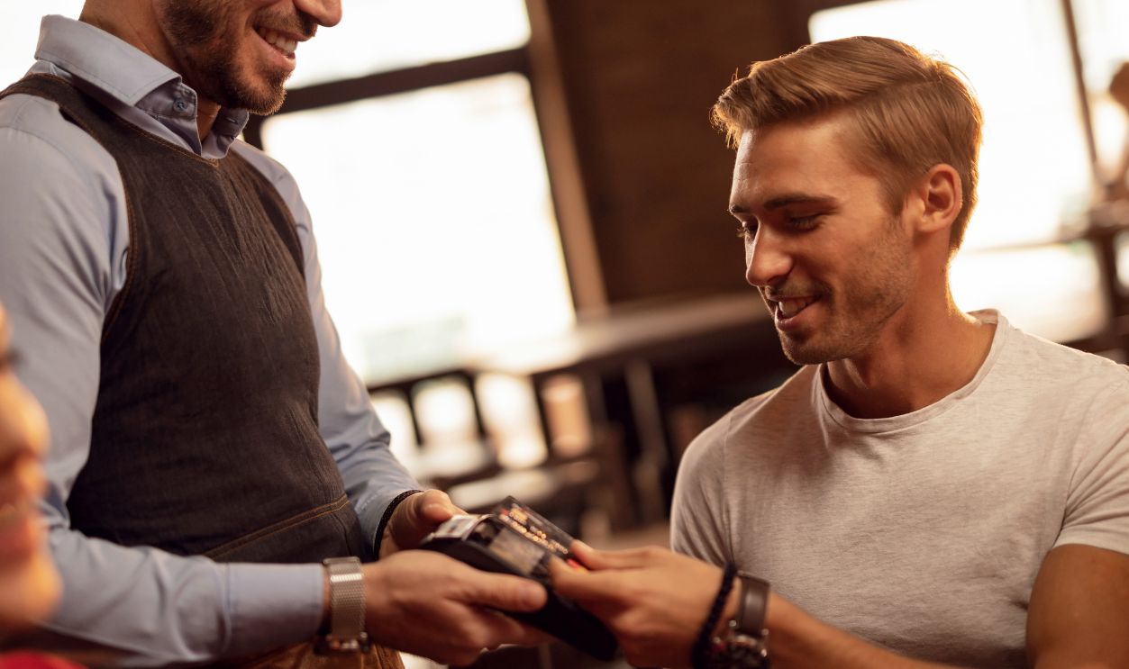 Young happy man paying waiter for restaurant check with contactless credit card.
