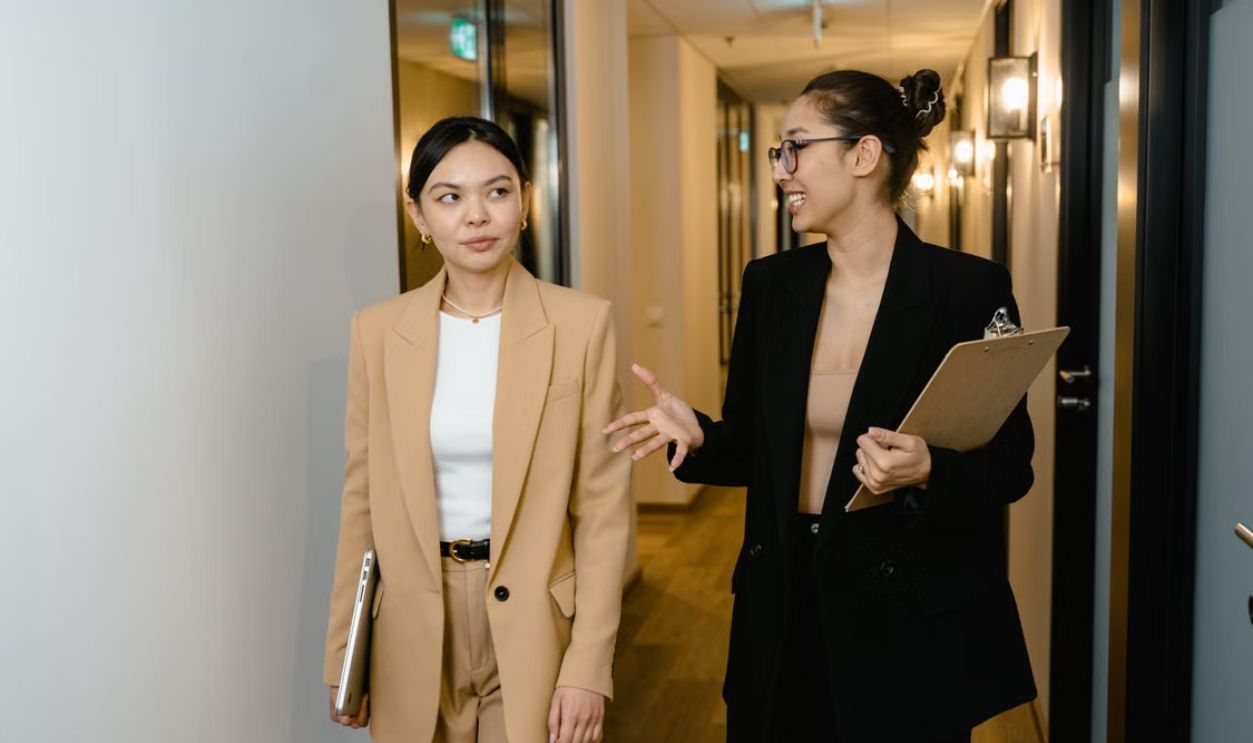 Two businesswomen converse while walking through a modern office hallway, holding documents.