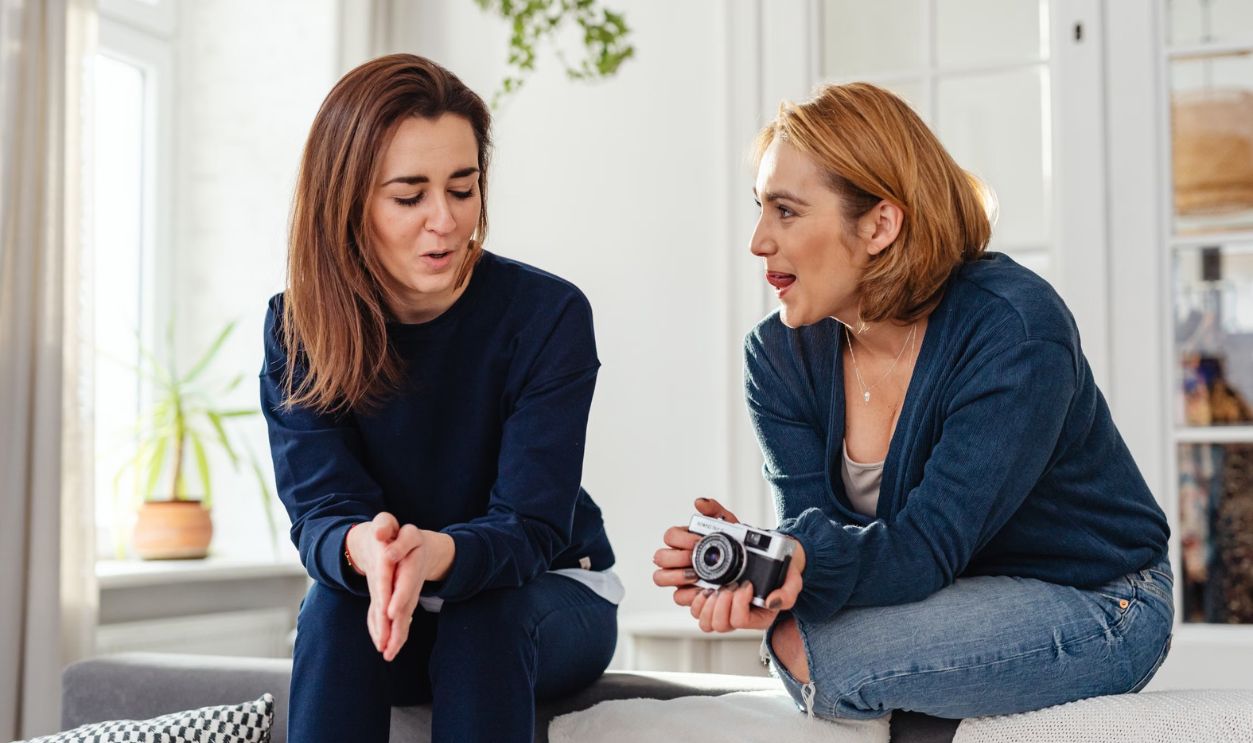 Two women in casual clothes sit on a couch, chatting and smiling warmly indoors.