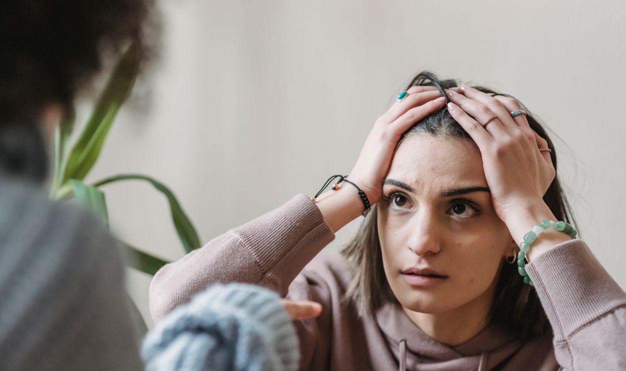 A woman in a beige hoodie looks distressed, holding her head with both hands during a conversation
