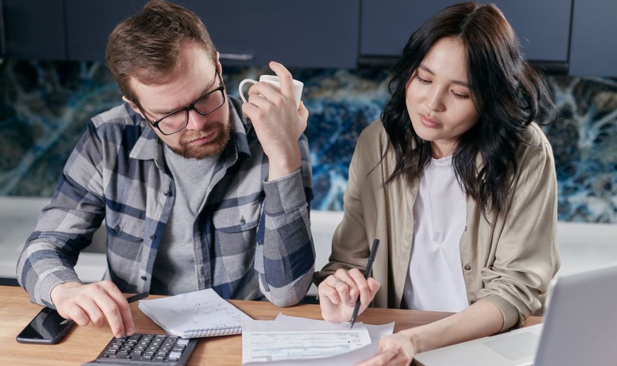 A couple reviewing household bills and budget using a calculator and laptop at their kitchen table.