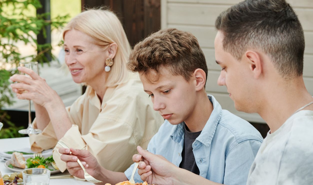 Mother and sons enjoying an outdoor meal with pasta and fresh fruits, captured in warm daylight.