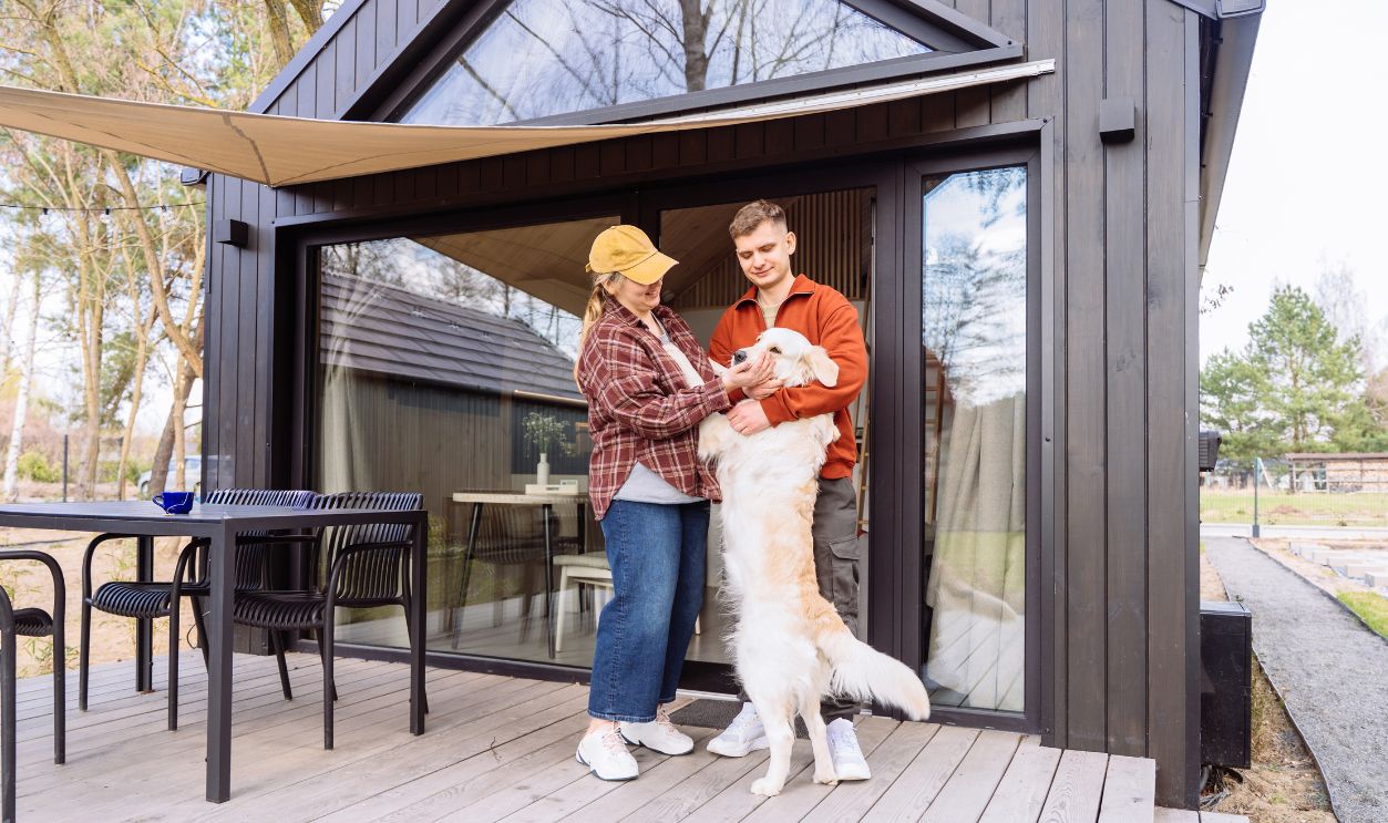 Smiling young couple enjoying playful moment with golden retriever near modern tiny house terrace. Cozy countryside vacation with pet friendly lifestyle, emotional connection and relaxing weekend mood.