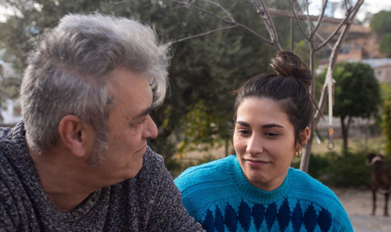 father and daughter talking outdoors with serious expression. gray-haired man in gray sweater in his fifties, twenty-something girl with bow tie and saturated blue sweater. in the background trees.