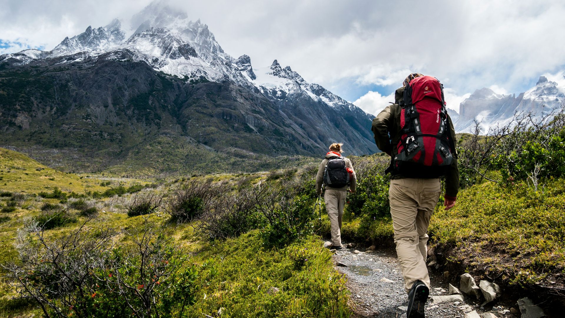 Two hikers with backpacks walking along a trail toward snow-capped mountains