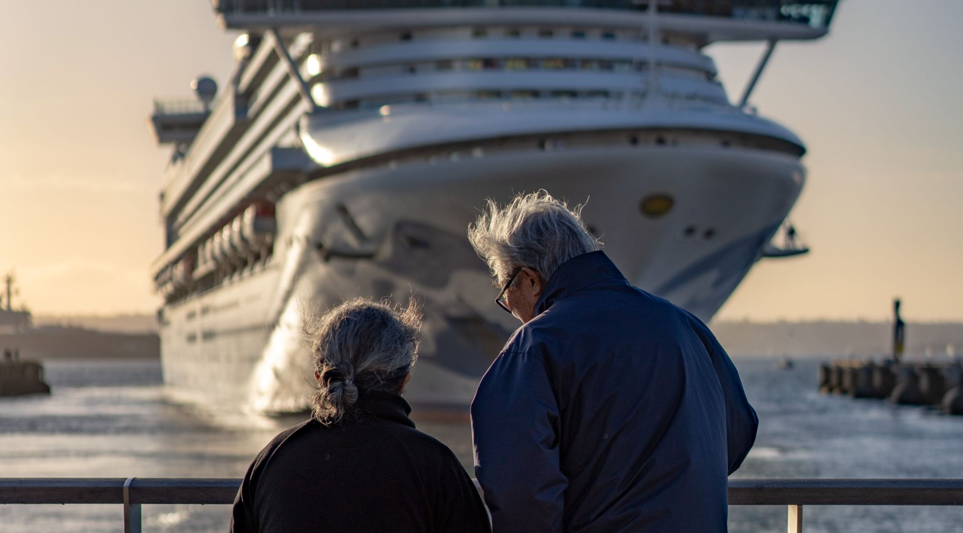 Senior couple standing at railing, admiring large cruise ship approaching dock at sunset.