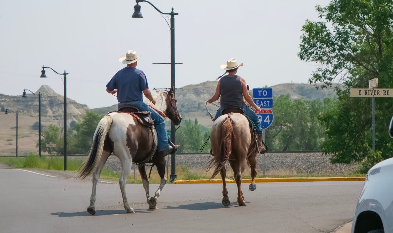MEDORA, NORTH DAKOTA — AUGUST 8, 2026: Two people in cowboy hats riding horses cross the asphalt road next to road signs and a car, showcasing the contrast of traditional transport and modern technology in the American West.