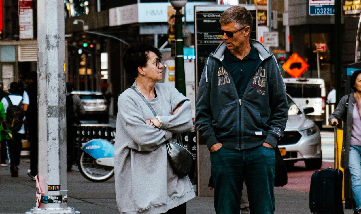 A man and woman stand at a crosswalk in New York City, surrounded by urban street life