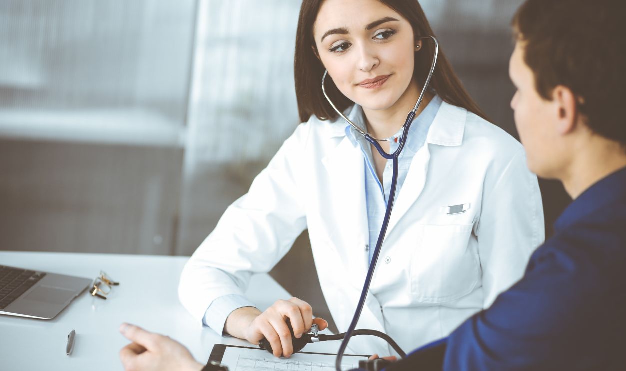 Young woman-doctor is checking her patient's blood pressure, while they are sitting together at the desk in a cabinet. Physician at work in a hospital. Medicine and healthcare concepts
