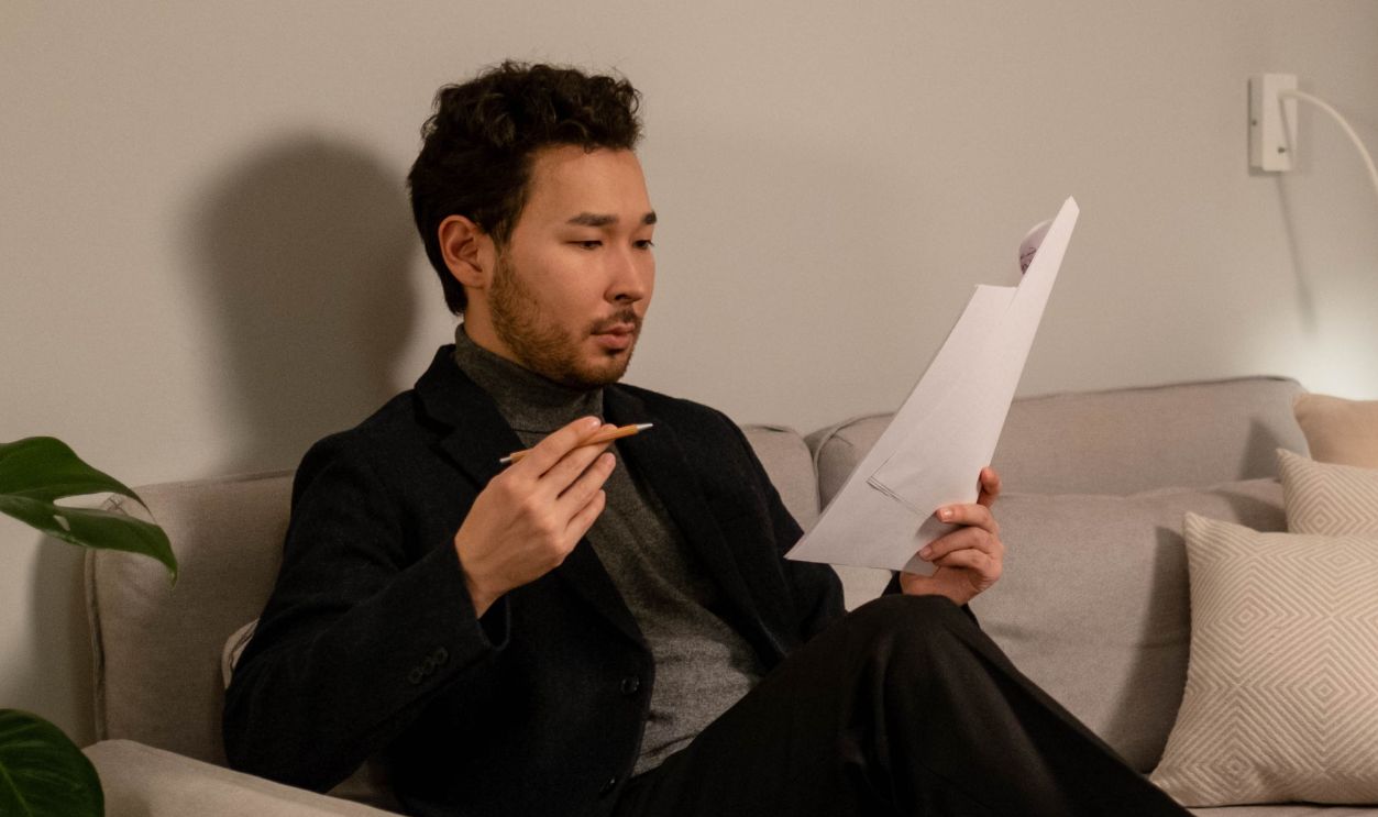 Asian man sitting on a couch reading documents, with a wine bottle on the table