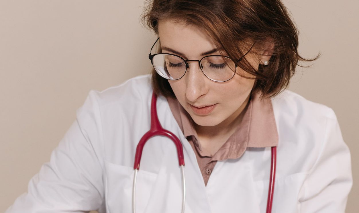 Female doctor with stethoscope writing notes in an office setting with laptop