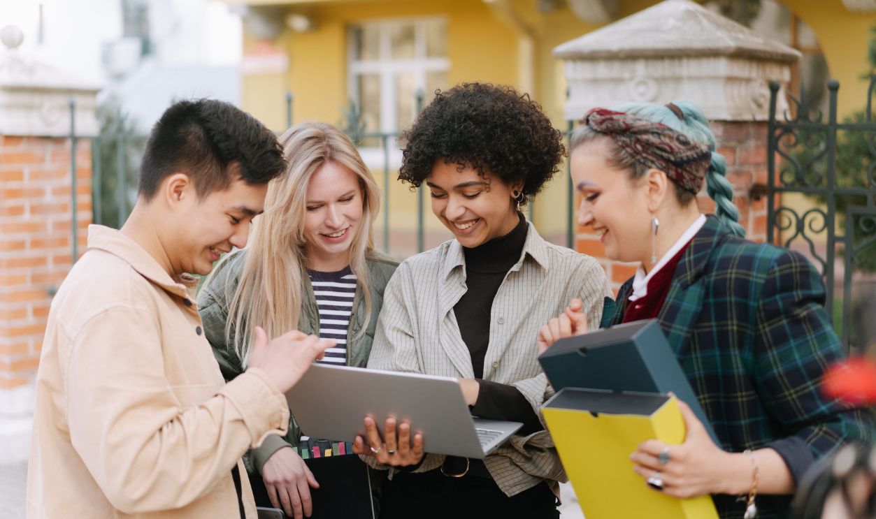A diverse group of young adults smiling and collaborating outdoors with a laptop