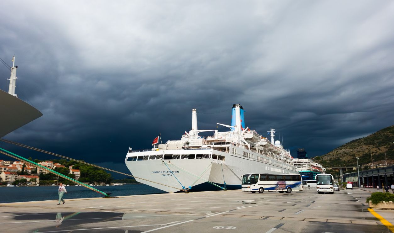 Shutterstock - 2797387643 - Marella Celebration cruise ship of Marella Cruises TUI during passenger embarkation, docked in Port of Dubrovnik Gruz under dramatic storm clouds, Adriatic sea.