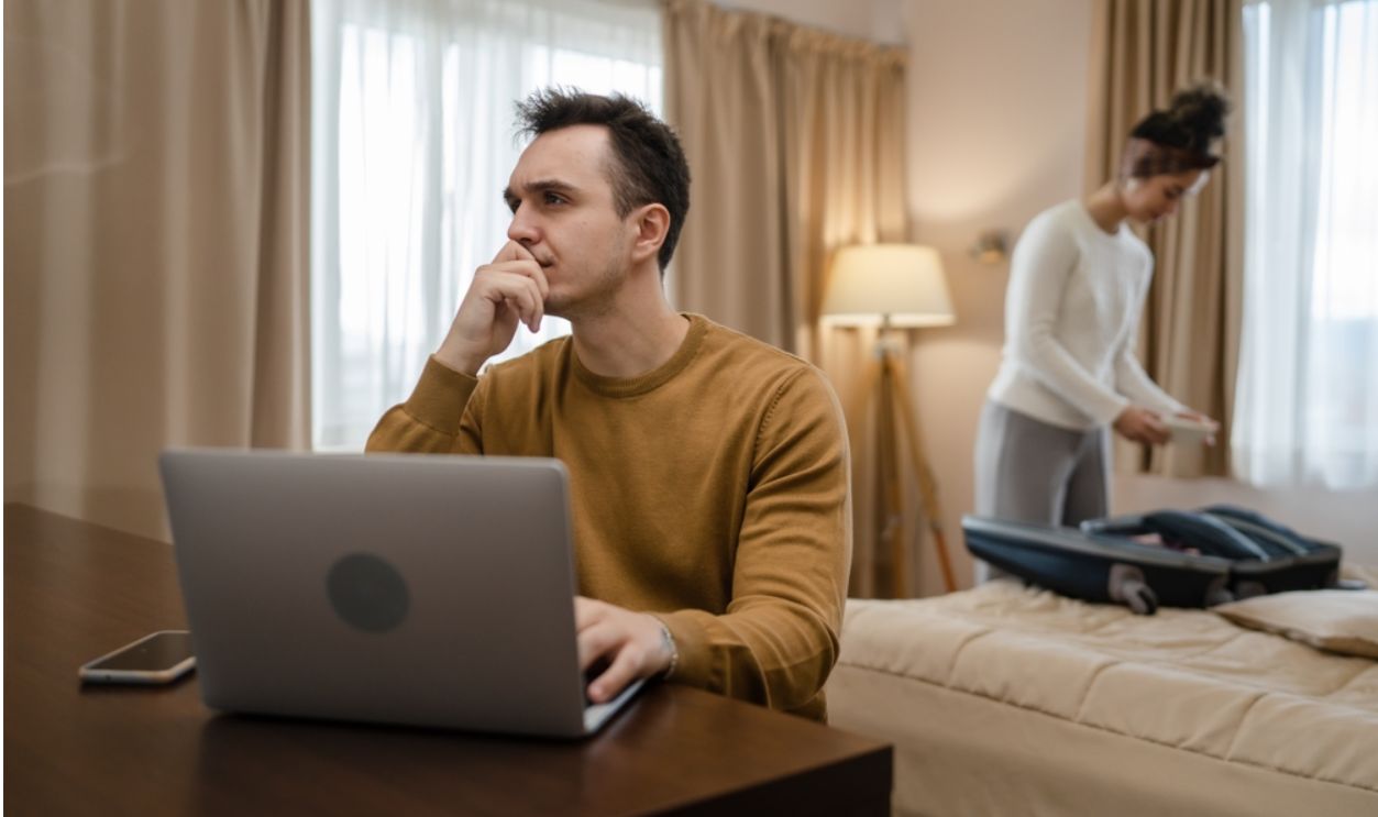 couple family life caucasian man working on laptop computer or making online reservations while his wife young woman is packing or unpack baggage suitcase on bed in hotel room real people copy space