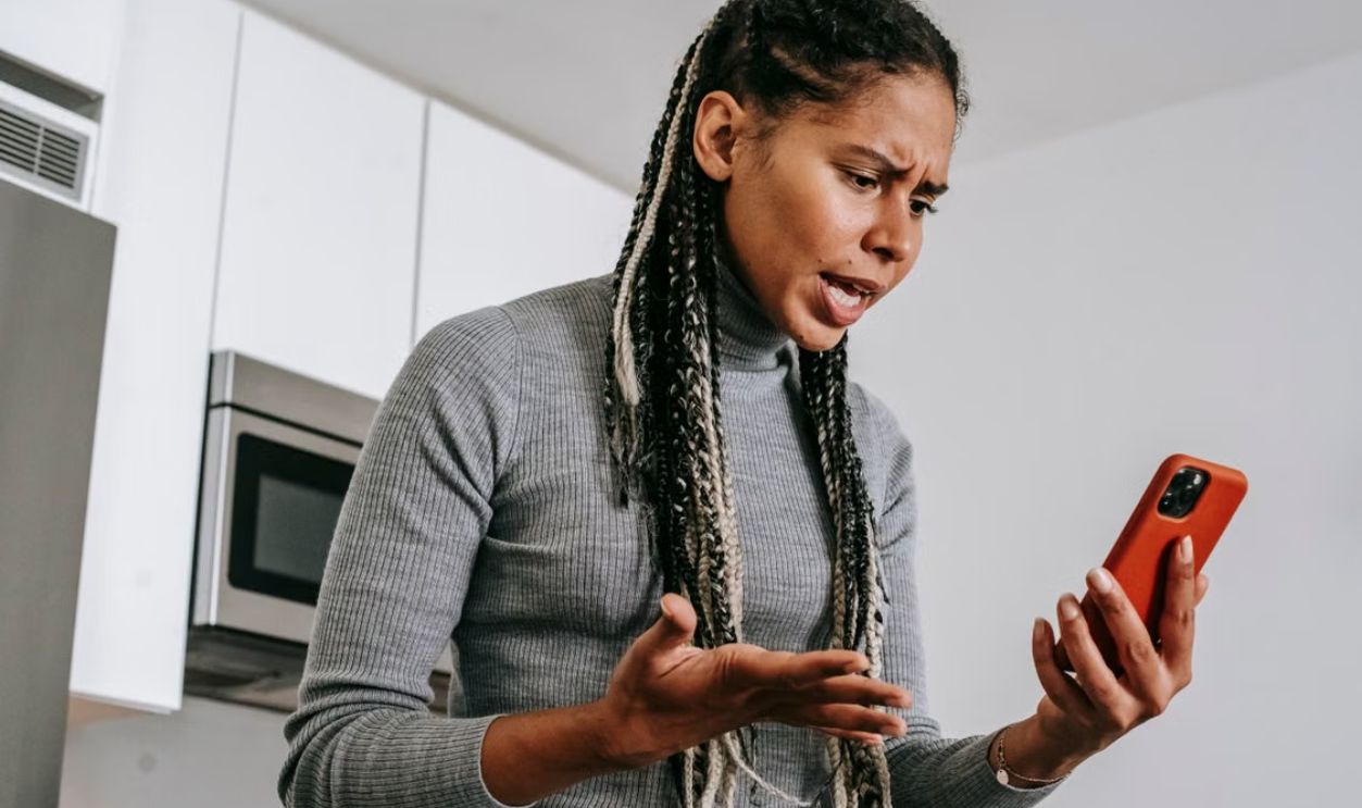 A young woman expressing frustration during a phone call in her kitchen, using a smartphone.