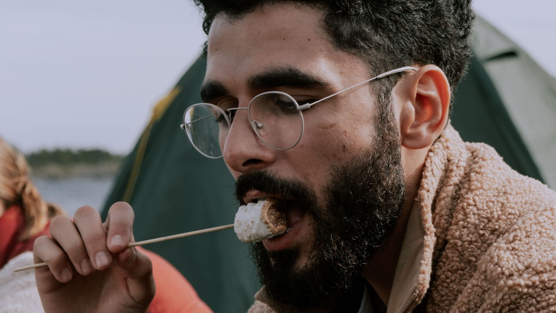 Bearded man enjoying marshmallow by campsite, embodying outdoor leisure.