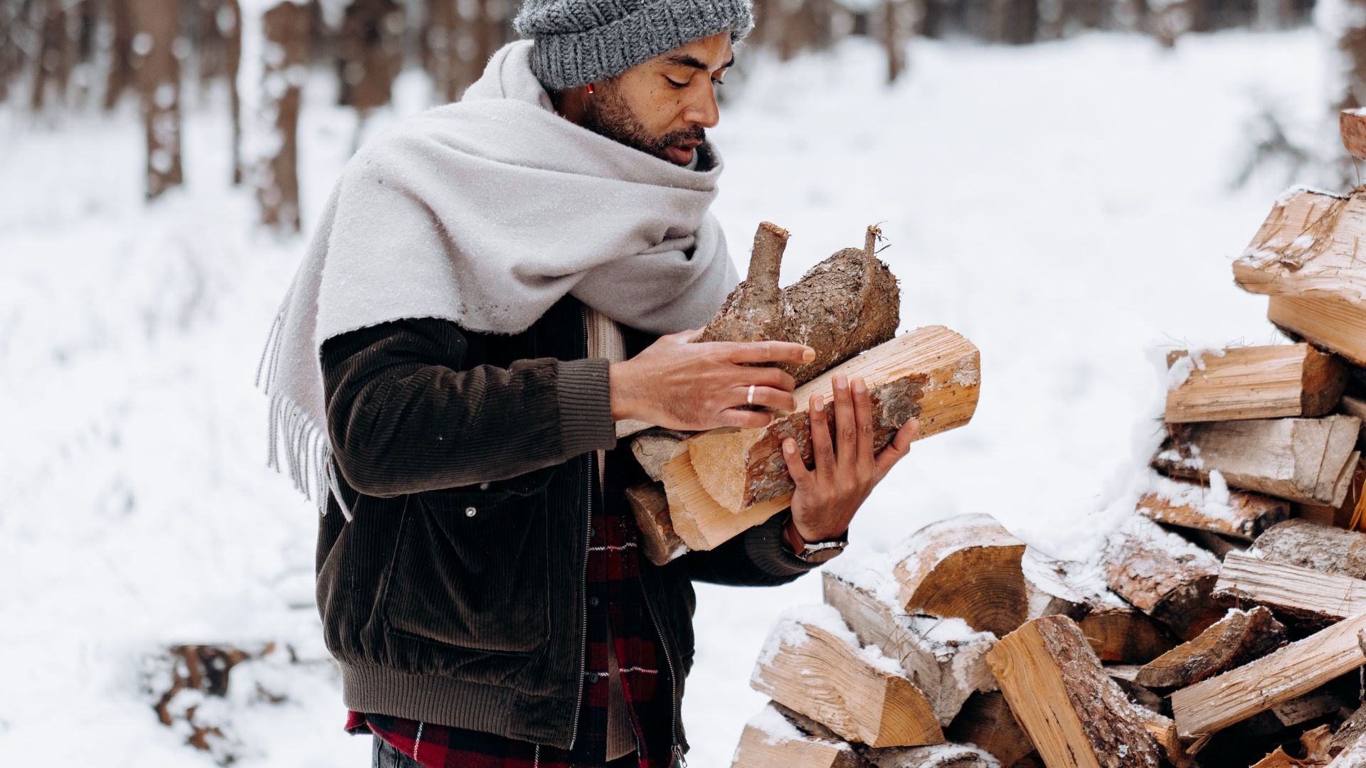 Person gathering logs in snowy winter forest, dressed in beanie and scarf, amidst tall trees.