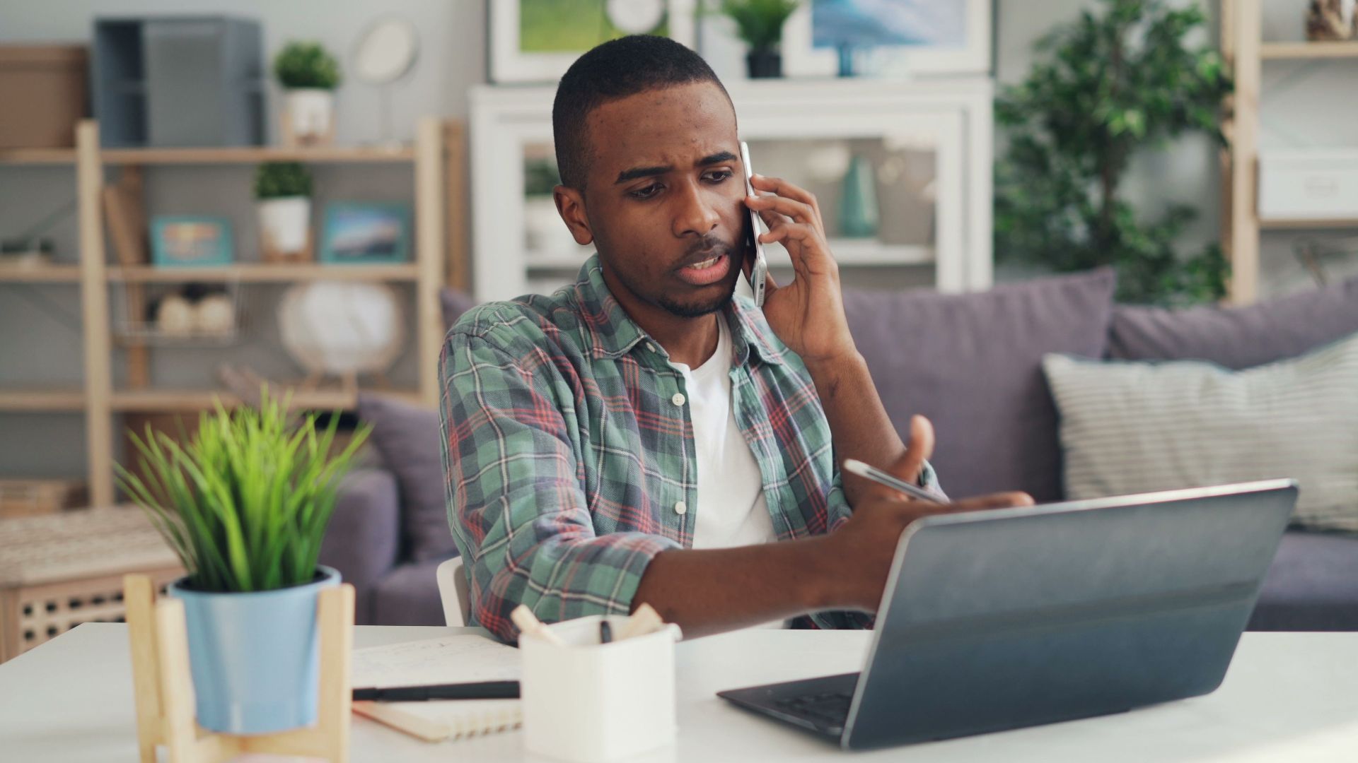Young man multitasking with phone and laptop in a cozy home office environment.