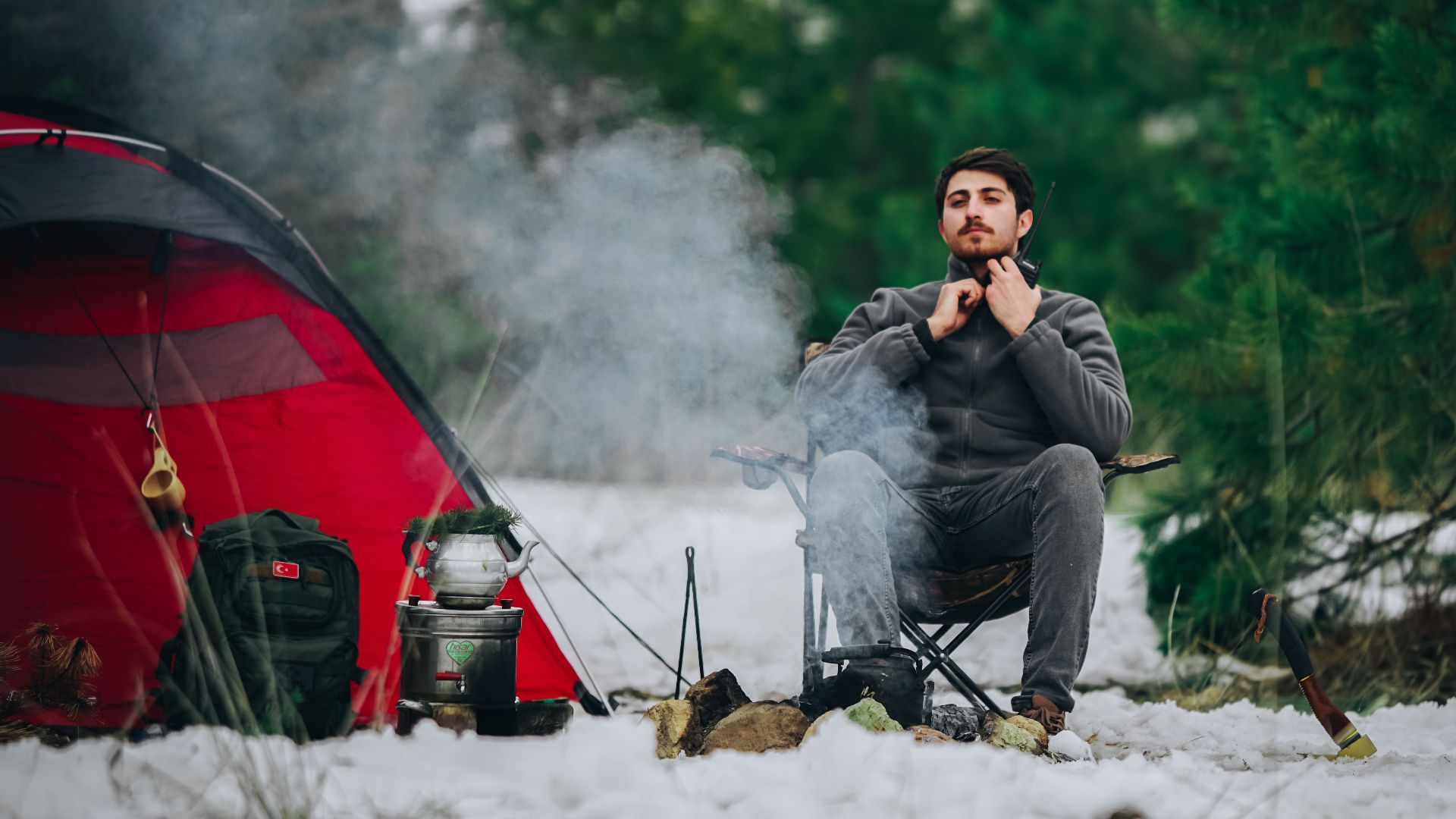 A man sitting by a red tent during a winter camping trip in a snowy forest.