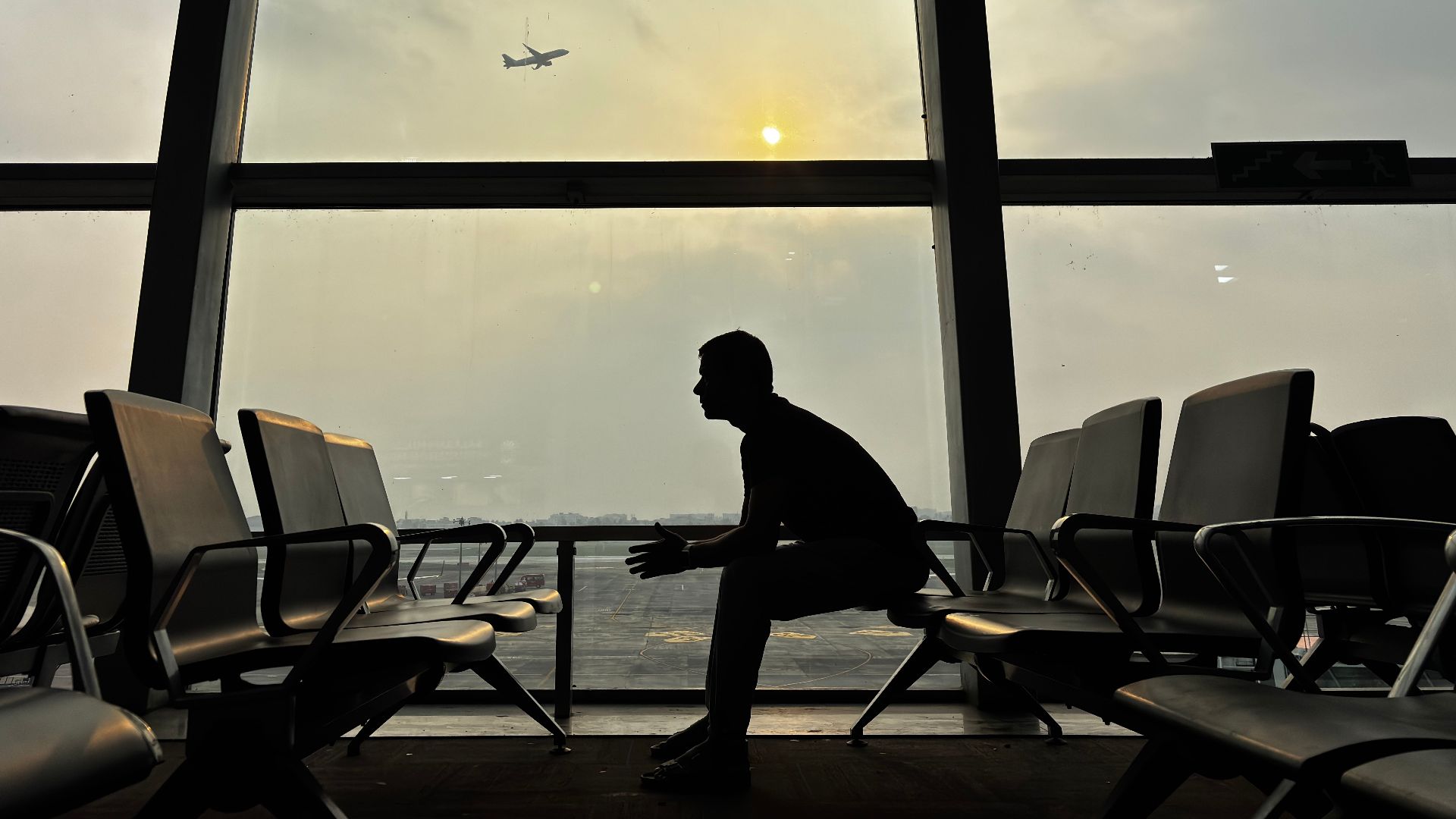 Silhouette of a man sitting at Kolkata Airport with a plane in the sky during sunset.