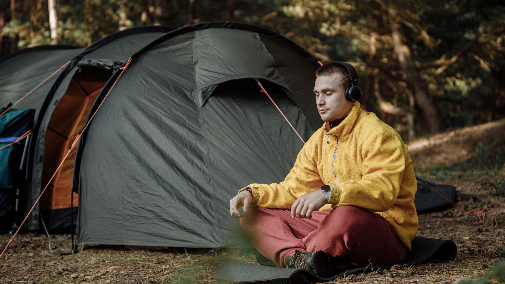 A man enjoys quiet meditation outside a tent in a forest, wearing headphones and a yellow jacket.