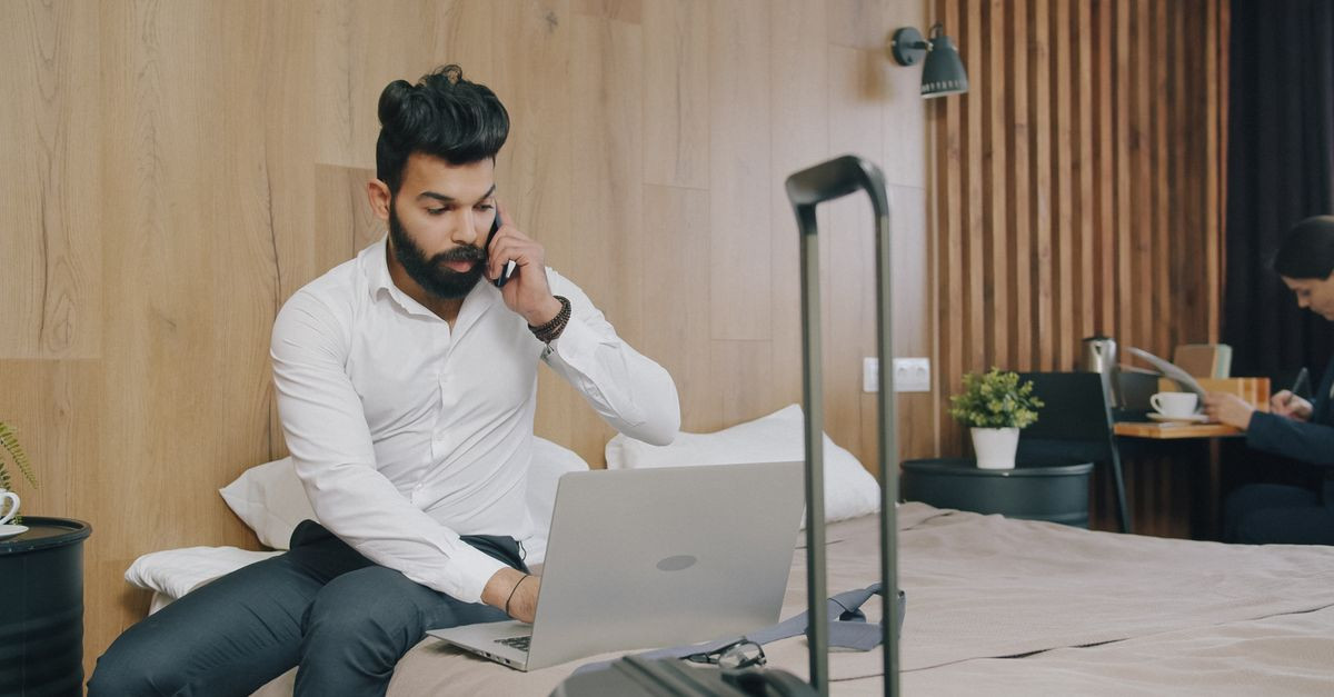 Businessman on laptop and phone in hotel room with luggage, working remotely.