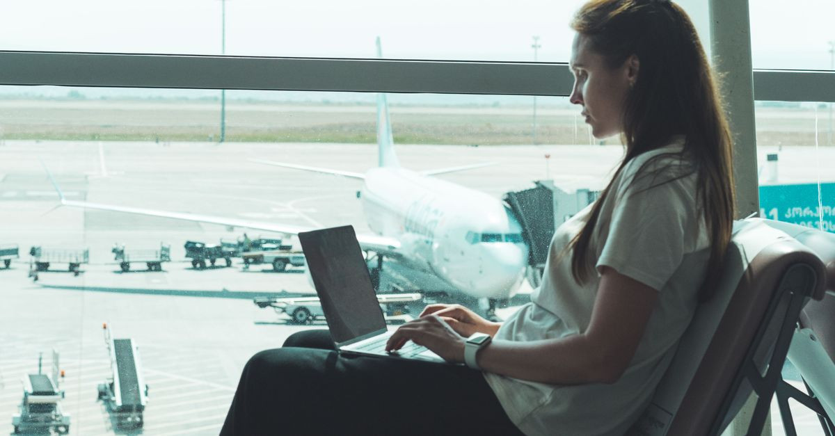 Side view of woman using laptop at airport, airplane visible outside through large glass windows.