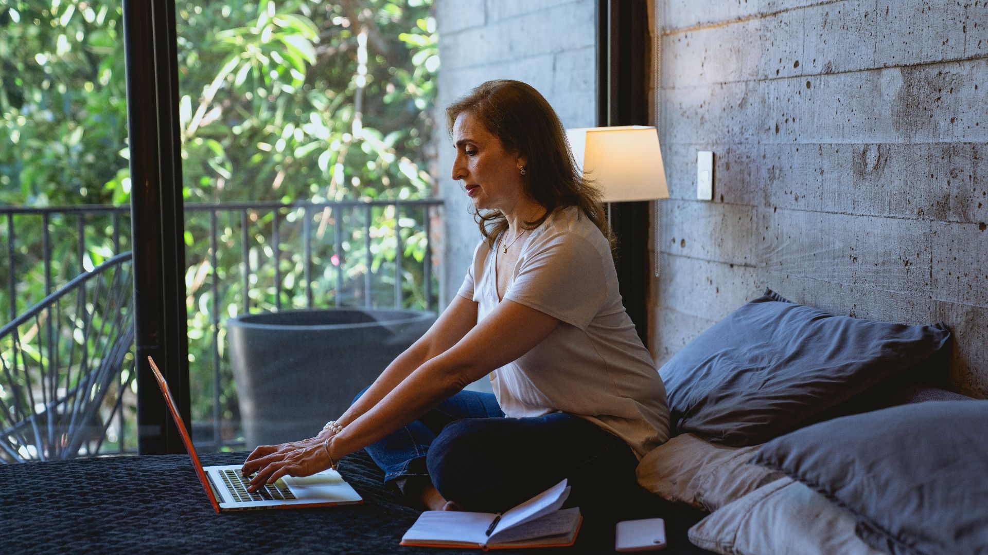 Senior Caucasian woman typing on a laptop in a cozy, modern bedroom with natural light.
