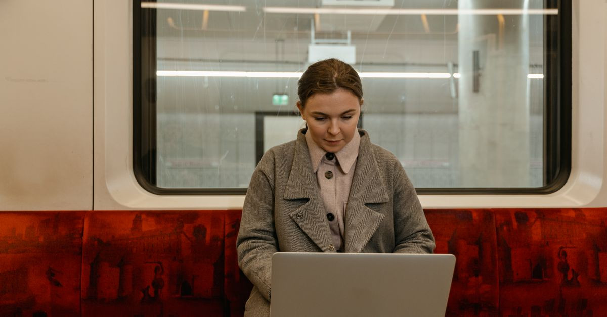Caucasian woman using laptop in a gray coat sitting inside a subway train.