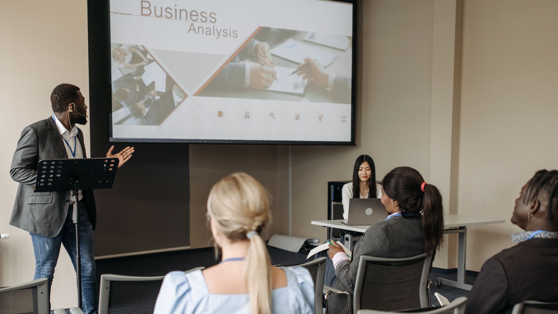 A diverse group attends a business analysis seminar with a presenter and projection screen in a modern conference room.