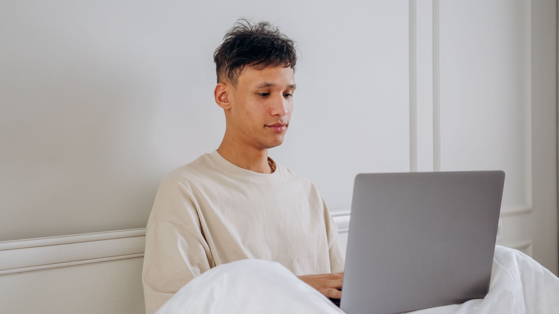 Young man sitting in bed using a laptop, embodying remote work lifestyle.
