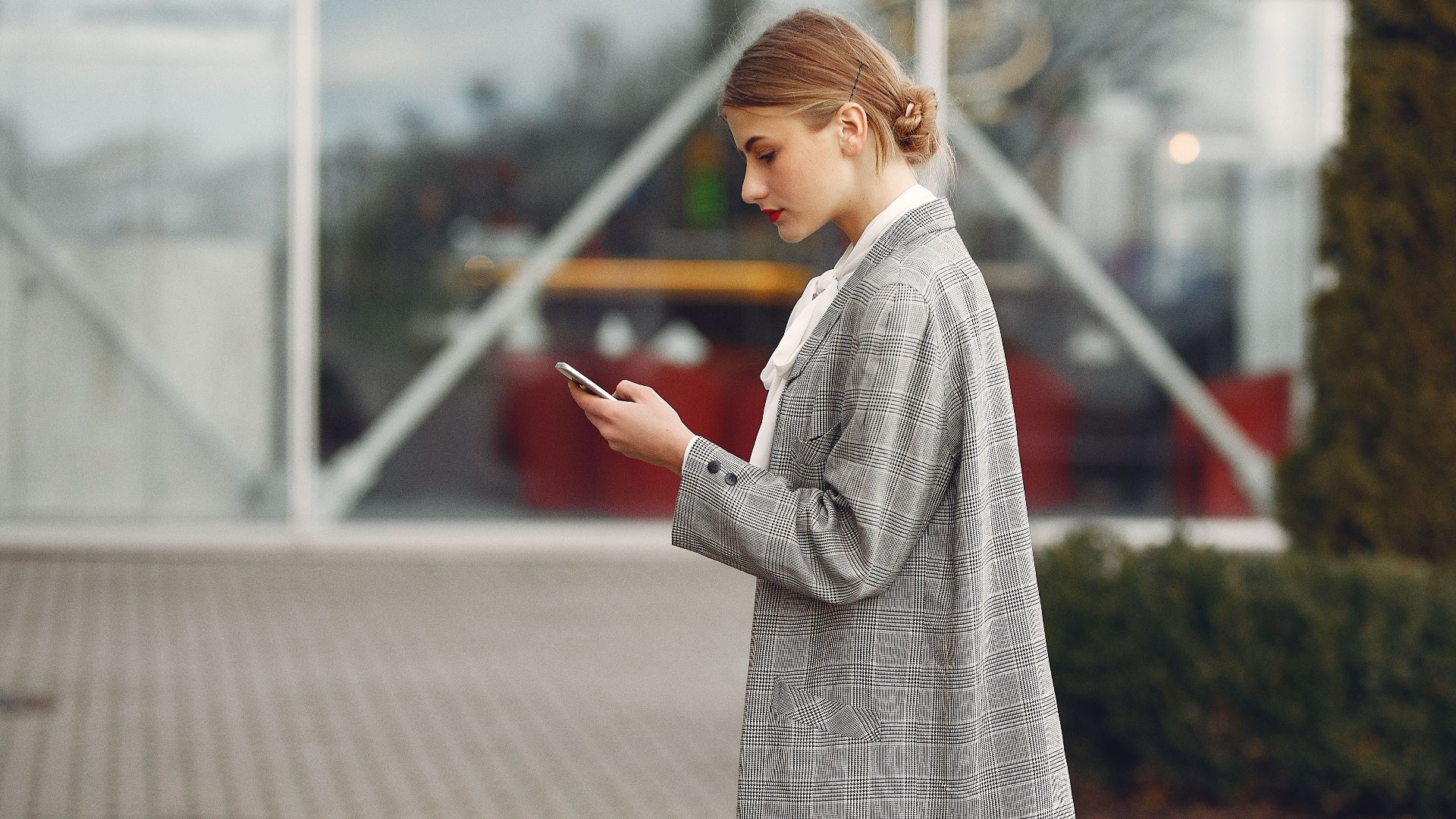 Stylish woman walking with suitcase while using smartphone outside a modern building.