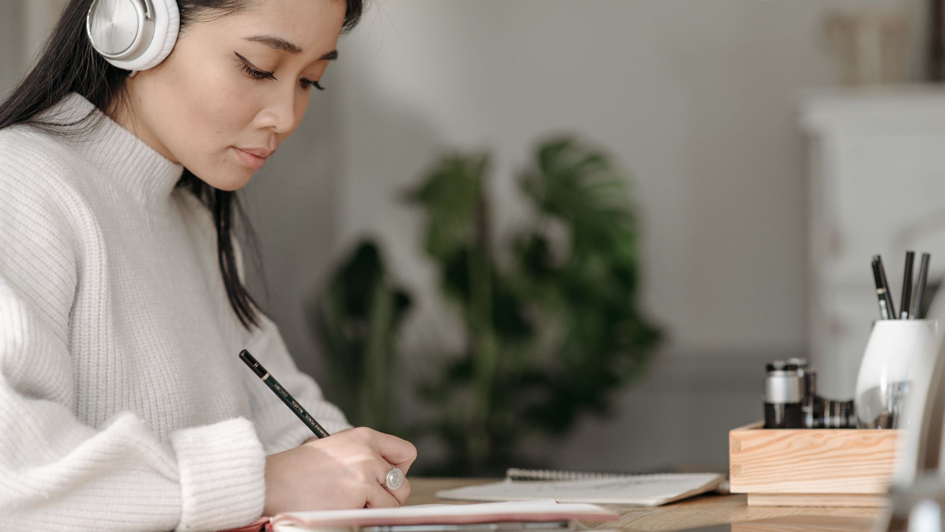 Asian woman wearing headphones writing at home, concentrating on her notes.