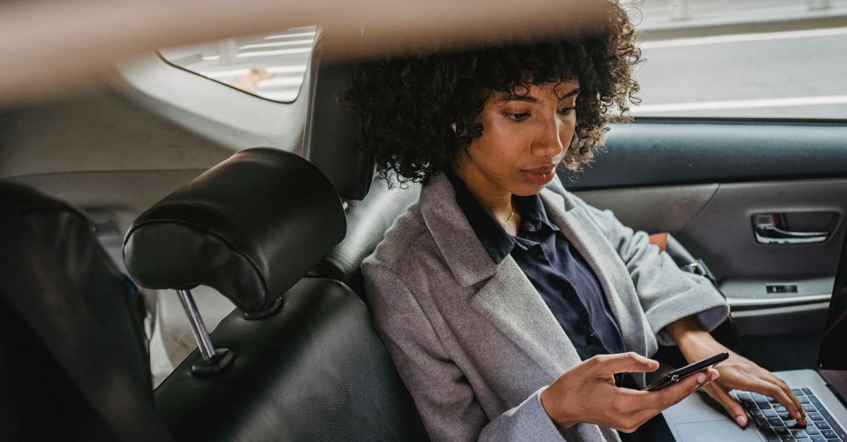 Woman multitasking with phone and laptop in car backseat, balancing work and travel.