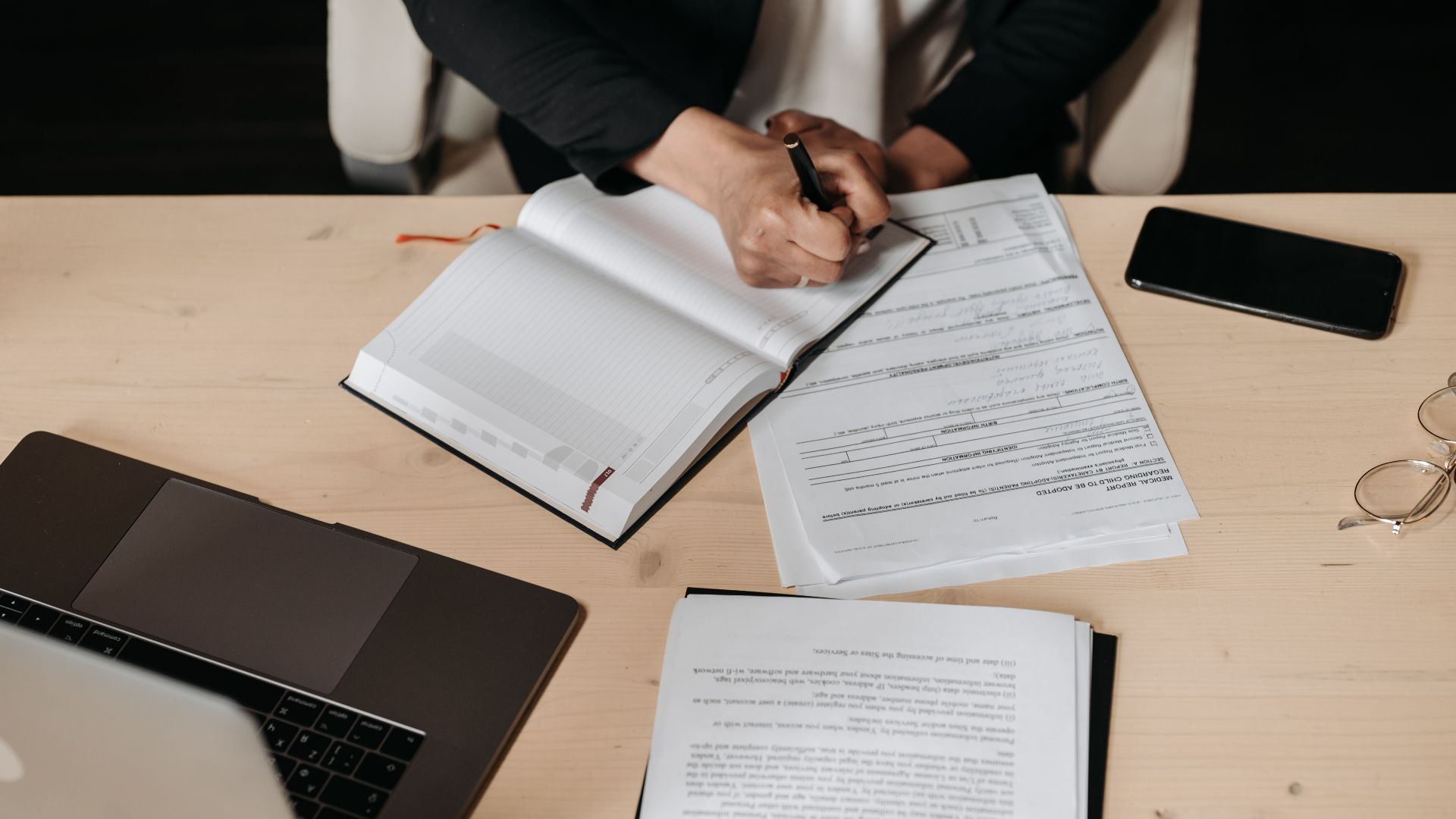 Overhead view of a desk setup with business documents, a laptop, and a person writing.