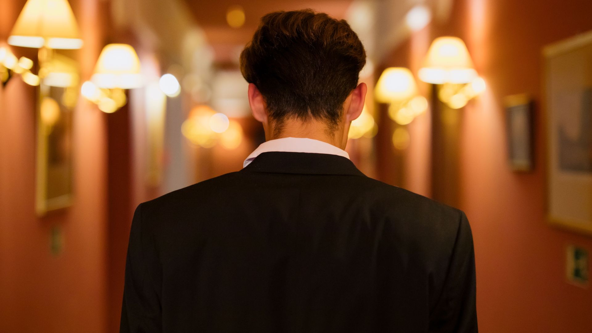 Rear view of man in black suit walking down a warmly lit hotel corridor.