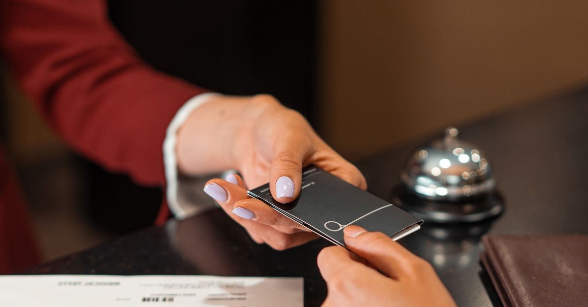 A hotel receptionist hands a key card to a guest over the counter, with documents visible.
