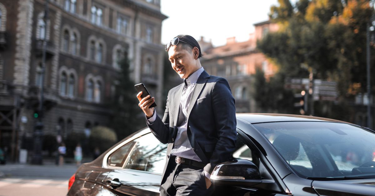 Smiling businessman in a black suit texting on his phone while leaning against a parked car in the city.