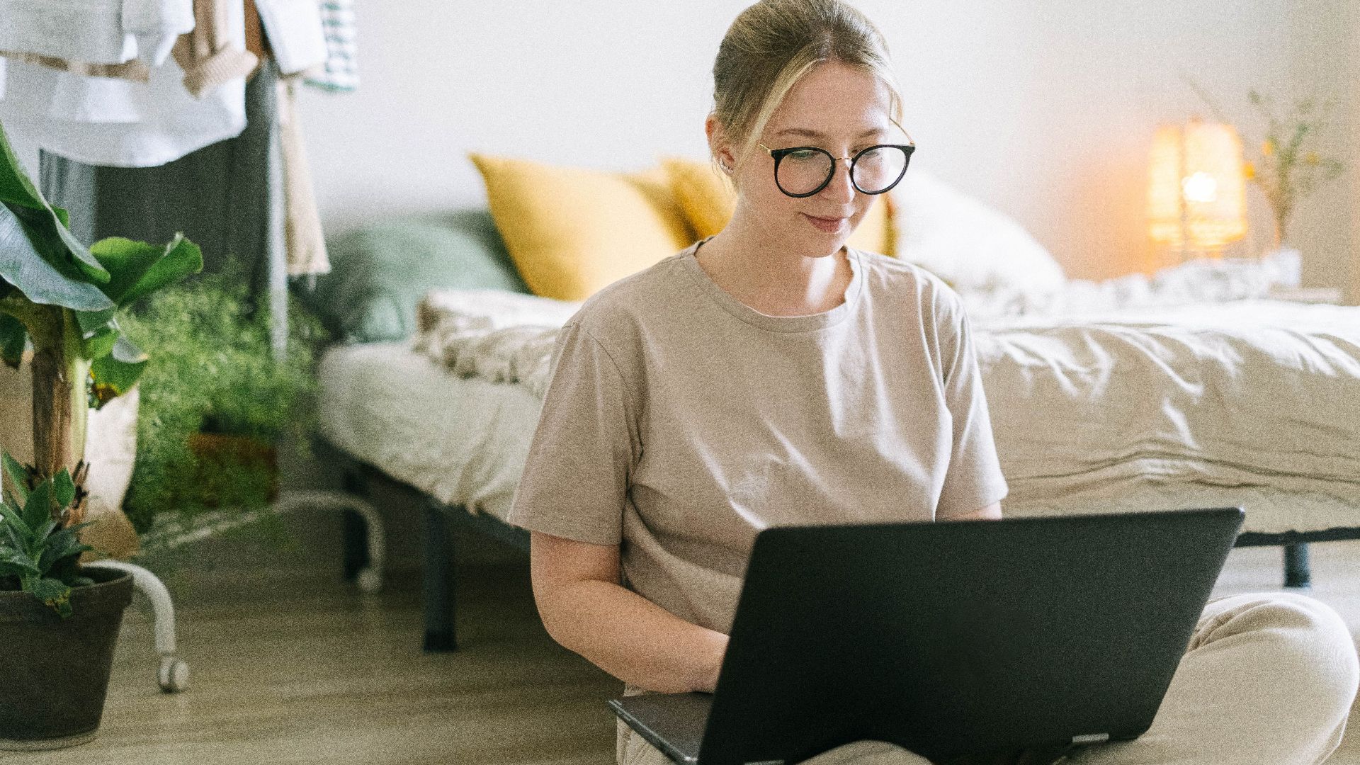 Caucasian woman wearing eyeglasses working on a laptop in a cozy bedroom setting.